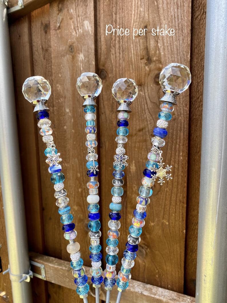 May include: Four decorative garden stakes with crystal ball tops and blue and white beads. The stakes have a snowflake charm at the bottom. The text "Price per stake" is visible at the top of the image.