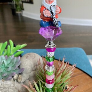 K&ouml;nnte beinhalten: Ein skurriler Gartenpfahl zeigt eine Gnome-Figur, die eine Schere h&auml;lt und auf einem violetten Kristall thront. Der Pfahl ist mit einer bunten Auswahl an Perlen in den Farben Rosa, Gr&uuml;n, Wei&szlig; und Silber verziert.