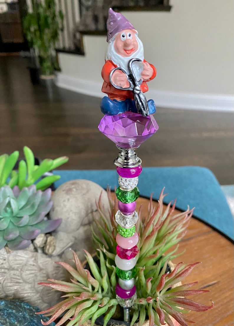 K&ouml;nnte beinhalten: Ein skurriler Gartenpfahl zeigt eine Gnome-Figur, die eine Schere h&auml;lt und auf einem violetten Kristall thront. Der Pfahl ist mit einer bunten Auswahl an Perlen in den Farben Rosa, Gr&uuml;n, Wei&szlig; und Silber verziert.