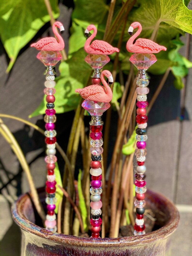 Pode incluir: Tr&ecirc;s estacas de jardim de flamingo rosa com detalhes de cristal e contas coloridas. As estacas s&atilde;o feitas de metal e t&ecirc;m uma variedade de contas em tons de rosa, vermelho, preto e branco.