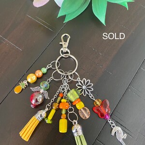 May include: A colourful keychain with a variety of charms, including a pineapple, a butterfly, a flower, and a bird. The keychain is made with beads, tassels, and metal charms. The keychain is sold.