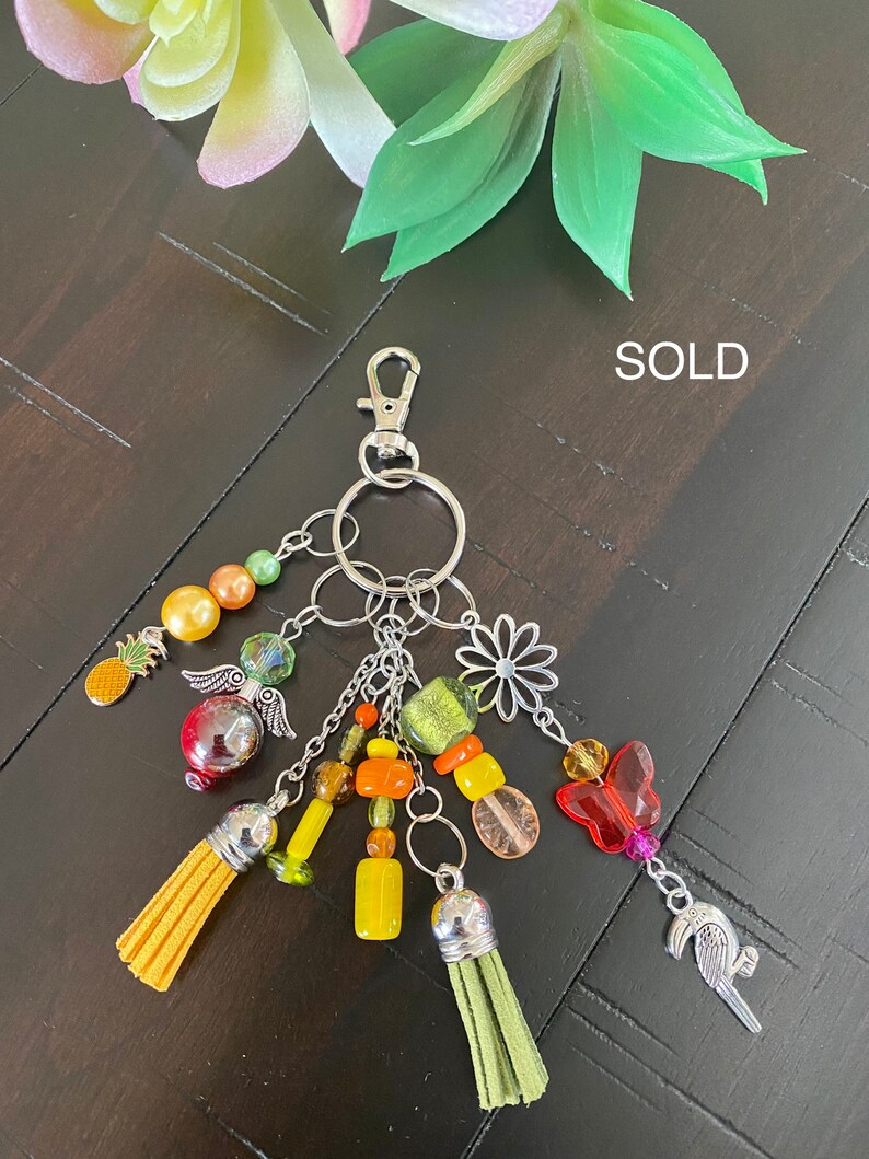 May include: A colourful keychain with a variety of charms, including a pineapple, a butterfly, a flower, and a bird. The keychain is made with beads, tassels, and metal charms. The keychain is sold.