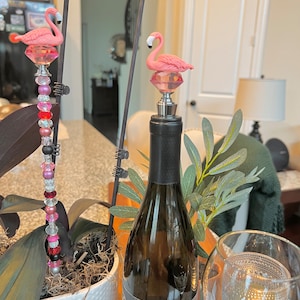 Pode incluir: Dois tamp&otilde;es de garrafa de flamingo rosa, um em uma garrafa de vinho e o outro em um caule decorativo com contas rosa e vermelhas.