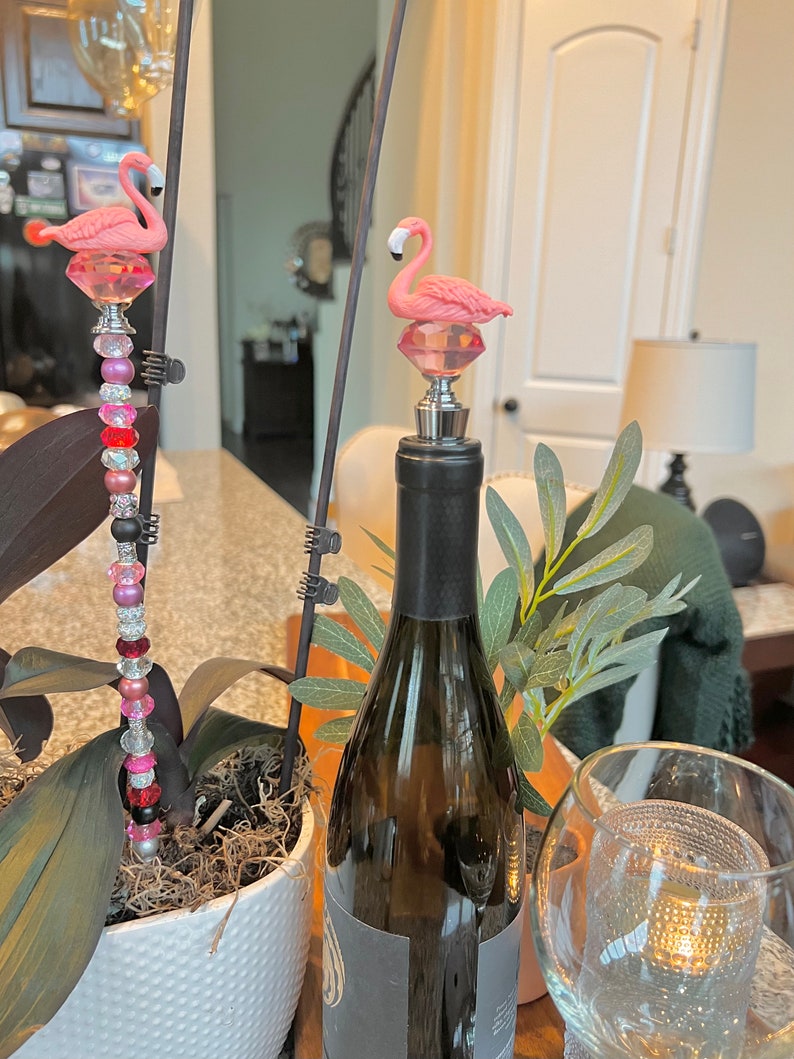 Pode incluir: Dois tamp&otilde;es de garrafa de flamingo rosa, um em uma garrafa de vinho e o outro em um caule decorativo com contas rosa e vermelhas.