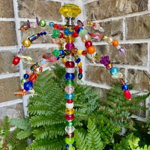 May include: A colourful beaded garden stake with a yellow crystal top. The stake is made of wire and is decorated with beads of various colours, including red, blue, green, and purple. The stake is surrounded by green ferns.