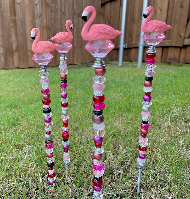 Pode incluir: Quatro estacas de jardim de flamingo rosa com hastes de contas. As estacas s&atilde;o decoradas com contas rosa, vermelhas, pretas e prateadas. Os flamingos est&atilde;o empoleirados em bases de cristal transparente.