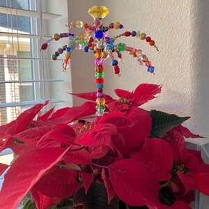 May include: A colourful beaded palm tree ornament with a yellow glass top, standing in a red poinsettia plant.