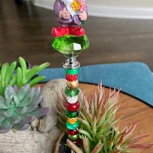 K&ouml;nnte beinhalten: Ein gr&uuml;ner, roter und goldener Perlenstab mit einer Kristallspitze. Eine kleine Gnomfigur mit einem lila Hemd und roten Schuhen befindet sich auf der Kristallspitze. Der Stab befindet sich in einem Topf mit einer gr&uuml;nen Sukkulente.