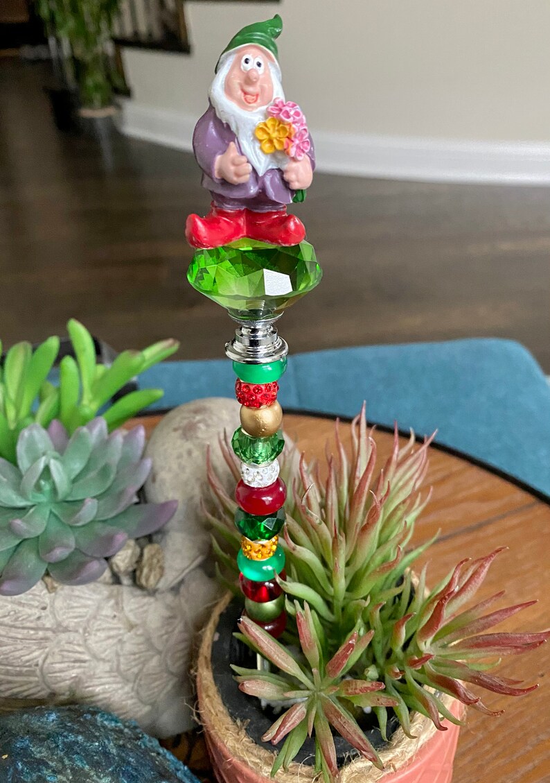 K&ouml;nnte beinhalten: Ein gr&uuml;ner, roter und goldener Perlenstab mit einer Kristallspitze. Eine kleine Gnomfigur mit einem lila Hemd und roten Schuhen befindet sich auf der Kristallspitze. Der Stab befindet sich in einem Topf mit einer gr&uuml;nen Sukkulente.