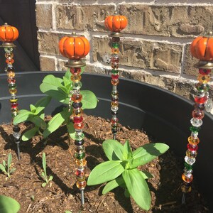 May include: Four orange pumpkin-shaped garden stakes with colorful glass beads. The stakes are inserted into a black planter with brown soil and green plants.