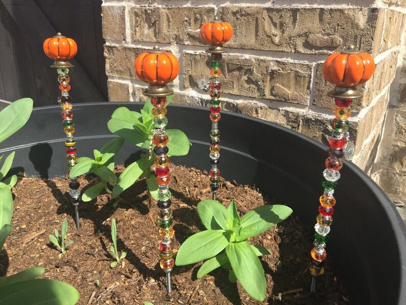 May include: Four orange pumpkin-shaped garden stakes with colorful glass beads. The stakes are inserted into a black planter with brown soil and green plants.