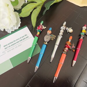 May include: Five colorful pens with decorative beads and charms. The pens are on a dark wood surface with a green card that says "When writing the story of your life, don't let anyone else hold your pen. Write well. Edit often!"