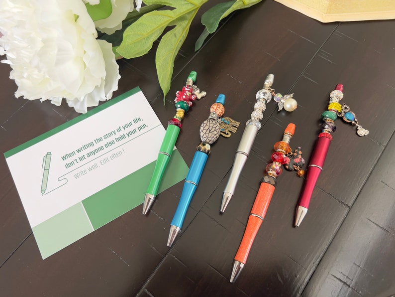 May include: Five colorful pens with decorative beads and charms. The pens are on a dark wood surface with a green card that says "When writing the story of your life, don't let anyone else hold your pen. Write well. Edit often!"