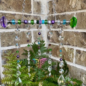 May include: A crystal suncatcher with a rainbow dragonfly charm hangs from a silver chain. The suncatcher features a beaded bar with green, purple, and silver beads. The crystal drops are clear and iridescent.
