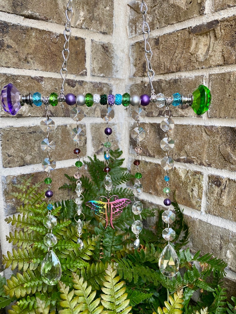 May include: A crystal suncatcher with a rainbow dragonfly charm hangs from a silver chain. The suncatcher features a beaded bar with green, purple, and silver beads. The crystal drops are clear and iridescent.