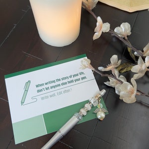 May include: A white card with green accents and the text "When writing the story of your life, don't let anyone else hold your pen. Write well, Edit often!" A silver pen with a pearl and angel wing charm is on the card.