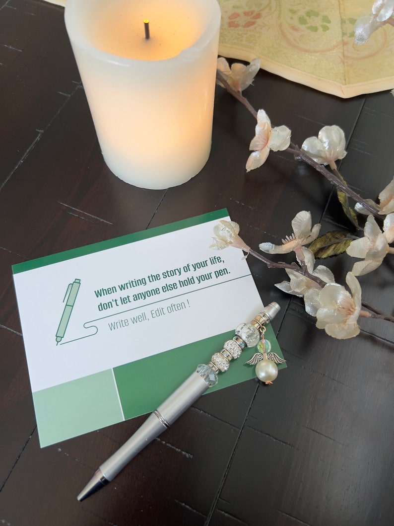 May include: A white card with green accents and the text "When writing the story of your life, don't let anyone else hold your pen. Write well, Edit often!" A silver pen with a pearl and angel wing charm is on the card.