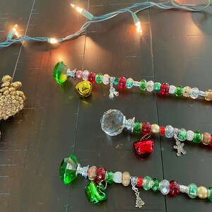 May include: Three beaded Christmas wands with crystal-shaped tops. Each wand has a different colored crystal: green, clear, and green. The wands are decorated with red, green, gold, and silver beads, and have various charms, including bells, a snowman, and a Christmas tree.