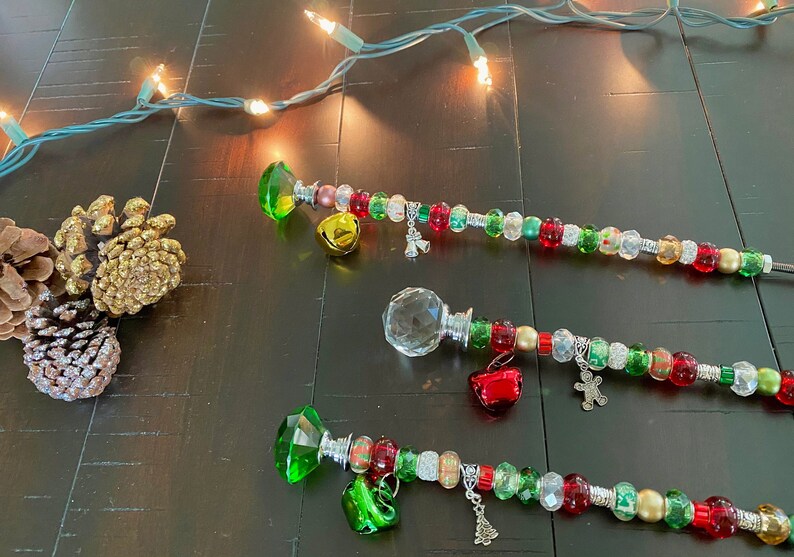 May include: Three beaded Christmas wands with crystal-shaped tops. Each wand has a different colored crystal: green, clear, and green. The wands are decorated with red, green, gold, and silver beads, and have various charms, including bells, a snowman, and a Christmas tree.