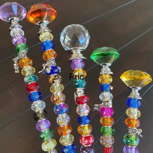 May include: Six colorful beaded garden stakes with crystal tops. The stakes are made of glass beads in various colors, including purple, orange, clear, green, and yellow. The tops of the stakes are crystal-shaped and have a silver finish. The text "Price is per stake" is visible at the top of the image.