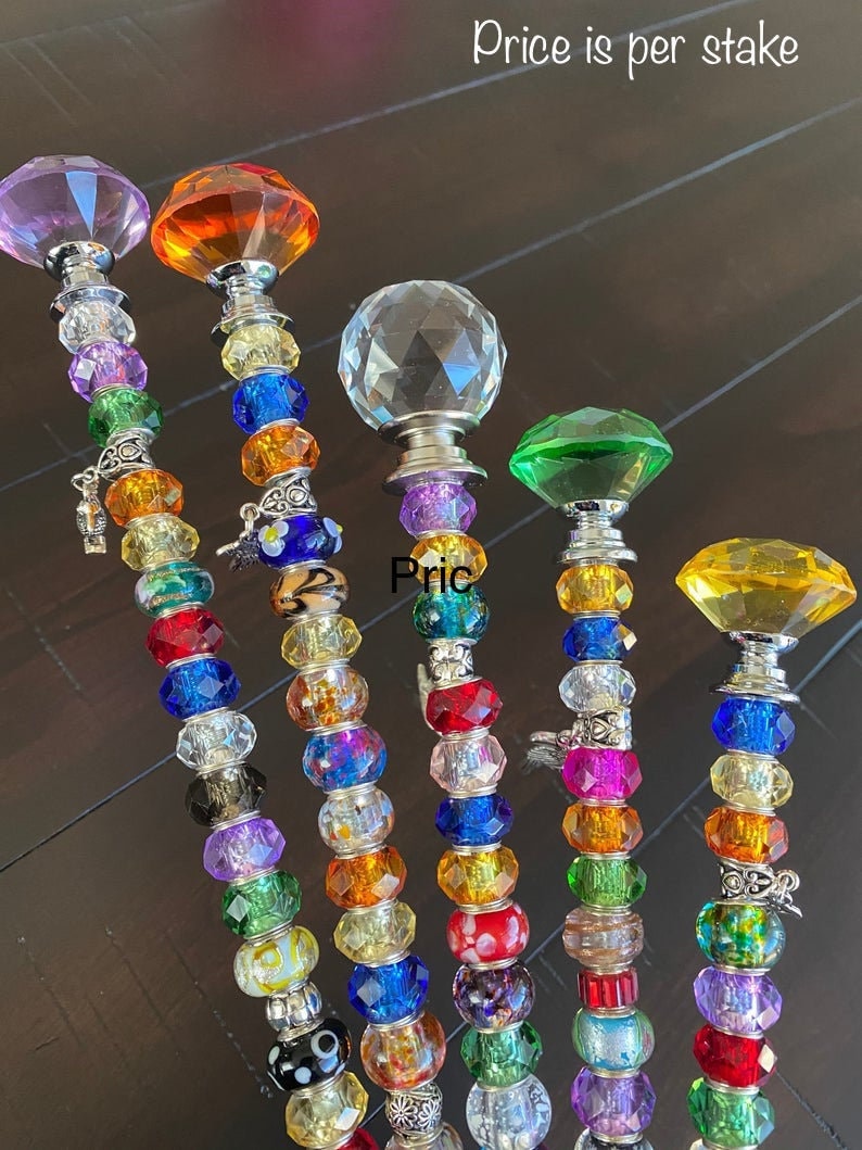 May include: Six colorful beaded garden stakes with crystal tops. The stakes are made of glass beads in various colors, including purple, orange, clear, green, and yellow. The tops of the stakes are crystal-shaped and have a silver finish. The text "Price is per stake" is visible at the top of the image.