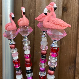 Pode incluir: Quatro estacas de jardim de flamingo rosa com topos de cristal e hastes de contas coloridas. As hastes de contas apresentam contas rosa, vermelhas, pretas, brancas e prateadas.