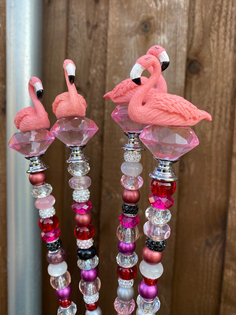 Pode incluir: Quatro estacas de jardim de flamingo rosa com topos de cristal e hastes de contas coloridas. As hastes de contas apresentam contas rosa, vermelhas, pretas, brancas e prateadas.