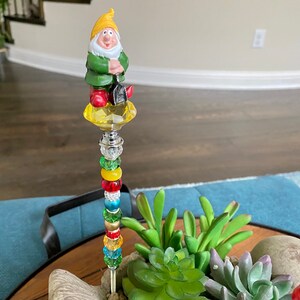 K&ouml;nnte beinhalten: Ein bunter Perlen-Gartenstab mit einer Gnome-Figur oben drauf. Der Gnome tr&auml;gt einen gr&uuml;nen Hut und h&auml;lt eine Schaufel. Der Stab ist mit roten, gelben, gr&uuml;nen und blauen Perlen verziert.