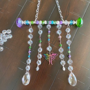May include: A crystal suncatcher with a rainbow butterfly charm and a beaded bar with green, purple, and blue beads. The suncatcher hangs from a silver chain.