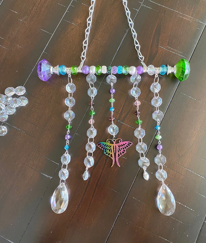 May include: A crystal suncatcher with a rainbow butterfly charm and a beaded bar with green, purple, and blue beads. The suncatcher hangs from a silver chain.