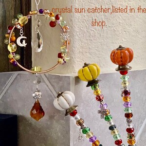 May include: Three beaded suncatchers with pumpkin-shaped ceramic accents. The suncatchers are made with colorful beads, including red, orange, yellow, green, purple, and clear. The beads are strung on wire and have a variety of shapes, including round, square, and star-shaped. The suncatchers are hanging from a white wall.