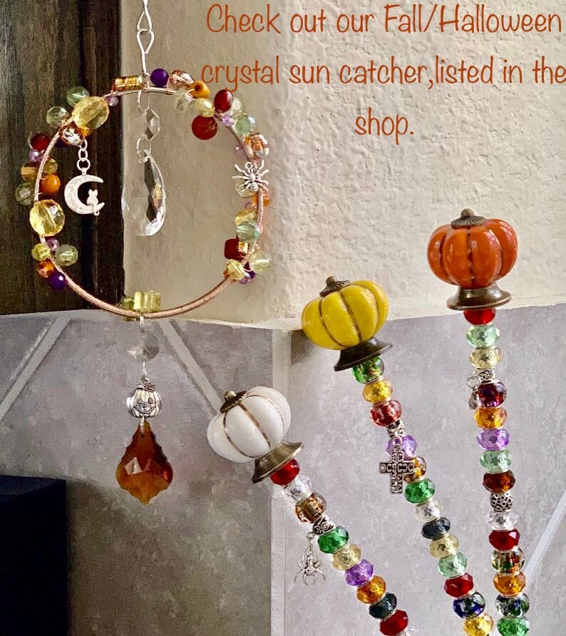 May include: Three beaded suncatchers with pumpkin-shaped ceramic accents. The suncatchers are made with colorful beads, including red, orange, yellow, green, purple, and clear. The beads are strung on wire and have a variety of shapes, including round, square, and star-shaped. The suncatchers are hanging from a white wall.