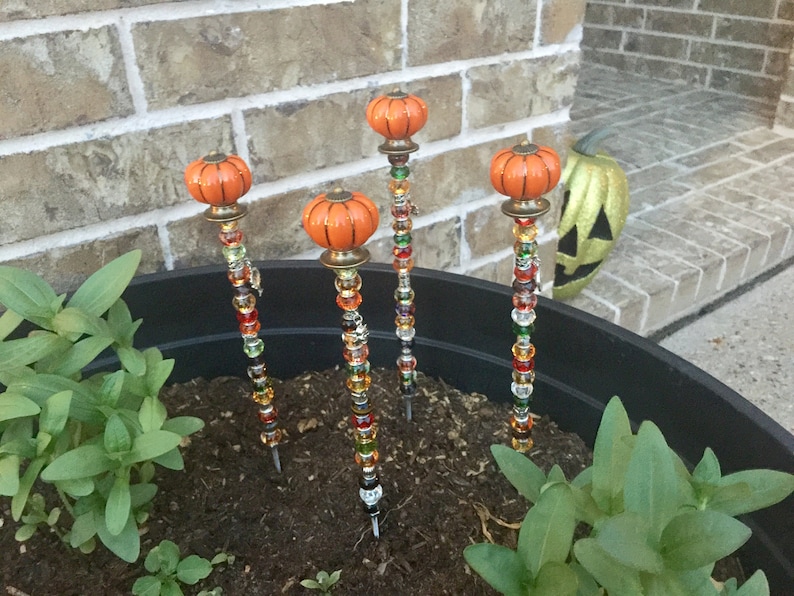 May include: Four decorative pumpkin-shaped garden stakes with colorful beads. The stakes are inserted into a black planter filled with soil and green plants.