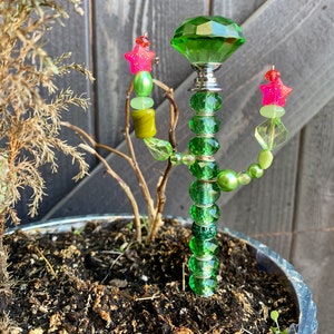 May include: A green glass cactus with a large green crystal on top. The cactus is decorated with green, yellow, and pink beads. The cactus is planted in a pot with brown soil.