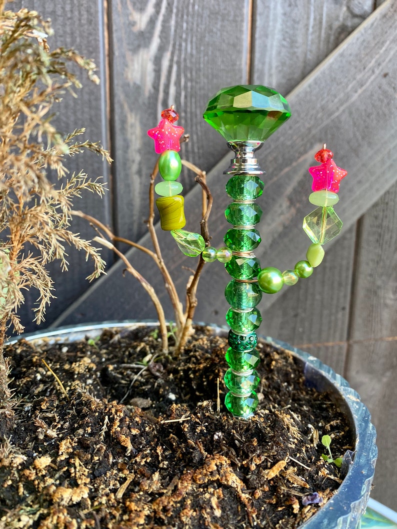 May include: A green glass cactus with a large green crystal on top. The cactus is decorated with green, yellow, and pink beads. The cactus is planted in a pot with brown soil.