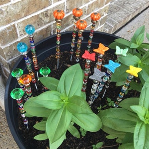 May include: A black planter pot filled with dark brown soil and a green plant.  The plant has large, green leaves.  There are 12 colorful garden stakes with butterfly and pumpkin-shaped toppers.  The stakes are made of beads and wire.