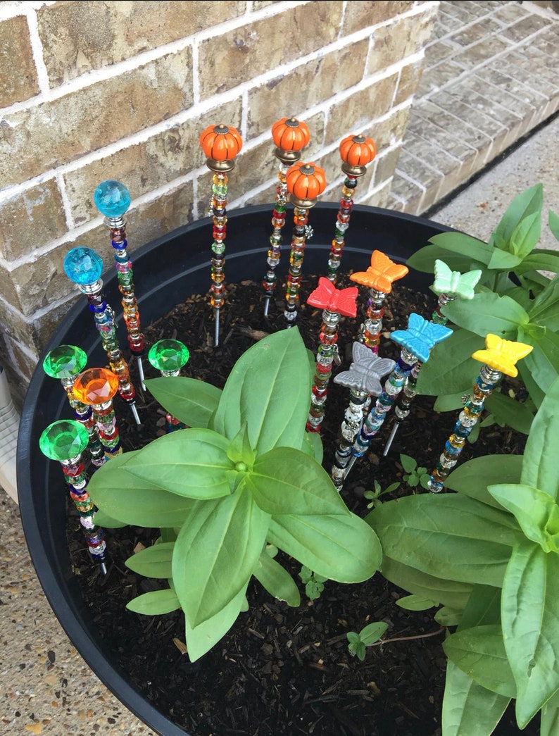 May include: A black planter pot filled with dark brown soil and a green plant.  The plant has large, green leaves.  There are 12 colorful garden stakes with butterfly and pumpkin-shaped toppers.  The stakes are made of beads and wire.