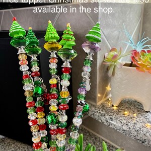 May include: Five beaded Christmas tree toppers with green, red, and white beads. The tree toppers are made of metal and have a small green Christmas tree on top. The text "Tree topper Christmas stakes are also available in the shop." is visible in the image.