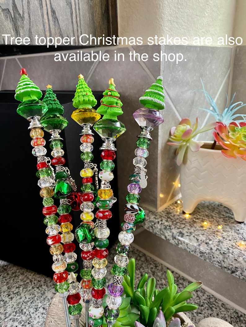 May include: Five beaded Christmas tree toppers with green, red, and white beads. The tree toppers are made of metal and have a small green Christmas tree on top. The text "Tree topper Christmas stakes are also available in the shop." is visible in the image.