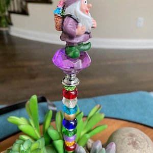 K&ouml;nnte beinhalten: Eine lilafarbene Zwergenfigur mit einem Blumenkorb auf dem R&uuml;cken steht auf einem lilafarbenen Kristall. Der Zwerg h&auml;lt eine Blume und hat einen wei&szlig;en Bart. Der Kristall befindet sich auf einem Stock mit bunten Perlen.