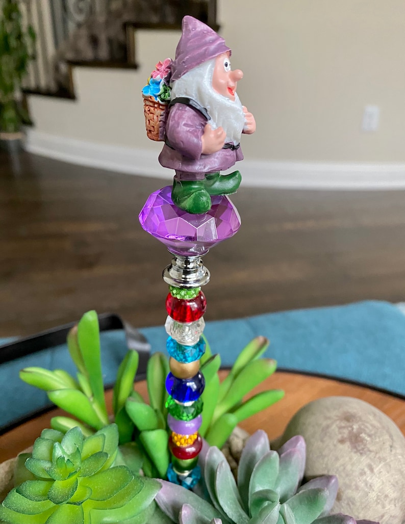 K&ouml;nnte beinhalten: Eine lilafarbene Zwergenfigur mit einem Blumenkorb auf dem R&uuml;cken steht auf einem lilafarbenen Kristall. Der Zwerg h&auml;lt eine Blume und hat einen wei&szlig;en Bart. Der Kristall befindet sich auf einem Stock mit bunten Perlen.