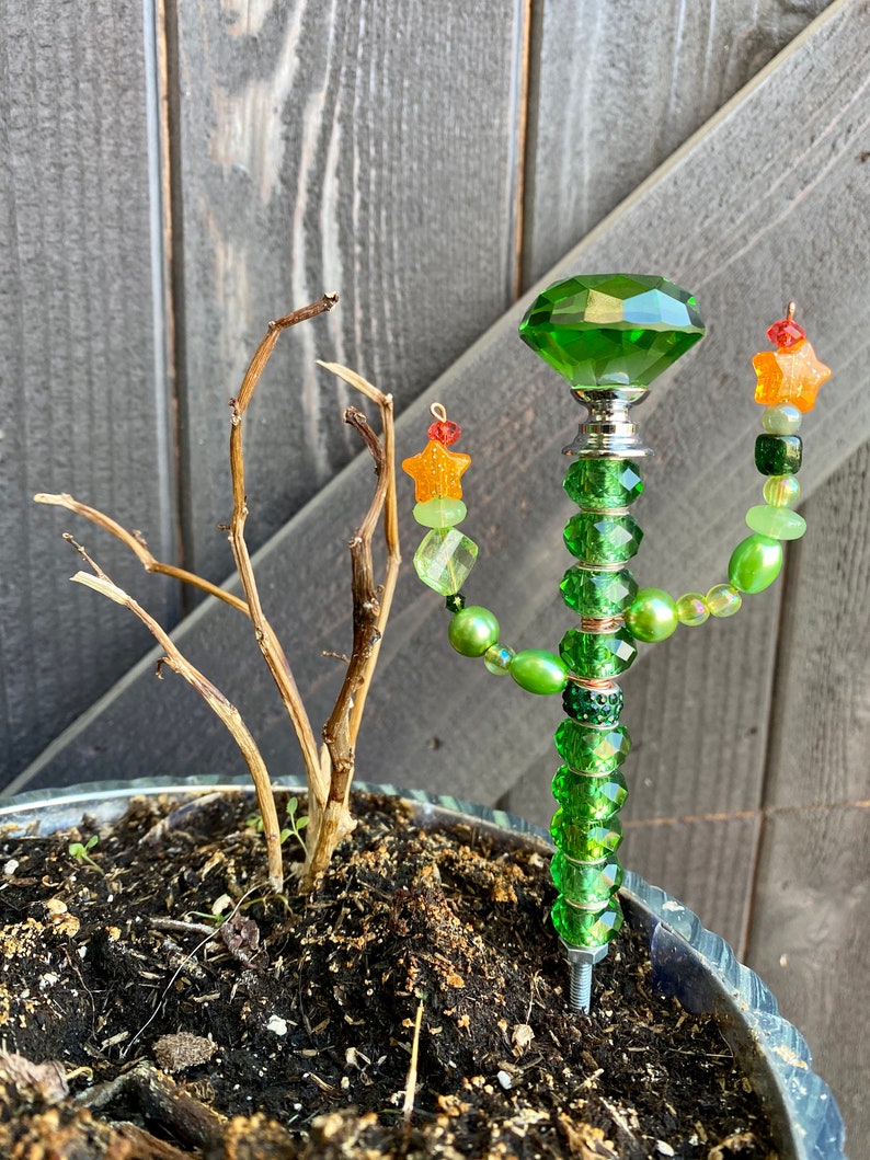 May include: A green glass beaded plant stake with a large green crystal at the top. The stake is decorated with green, orange, and gold beads and stars. The stake is inserted into a pot of soil.