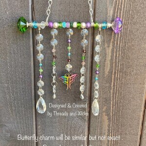 May include: A crystal suncatcher with a butterfly charm, featuring a rainbow of colors, including green, purple, and clear crystals. The suncatcher is hung from a silver chain and has a beaded bar with a pearl and green and purple crystal accents. "Designed & Created by Threads and Wicks" is written on the bottom of the suncatcher.