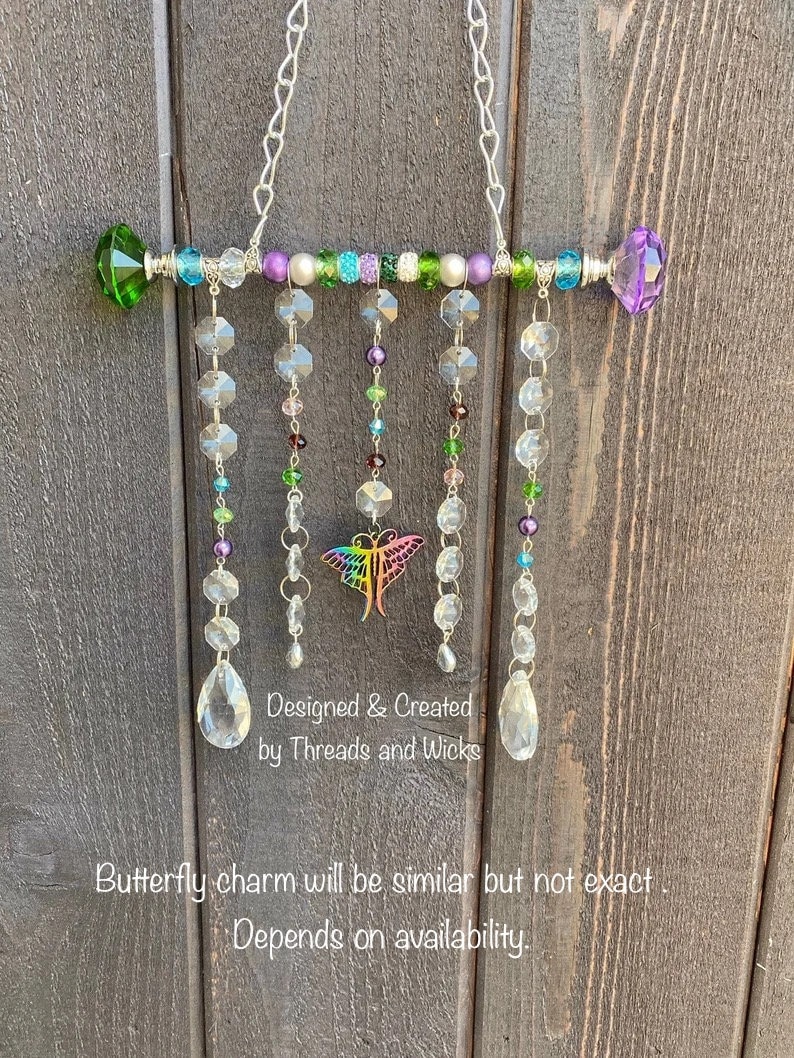 May include: A crystal suncatcher with a butterfly charm, featuring a rainbow of colors, including green, purple, and clear crystals. The suncatcher is hung from a silver chain and has a beaded bar with a pearl and green and purple crystal accents. "Designed & Created by Threads and Wicks" is written on the bottom of the suncatcher.