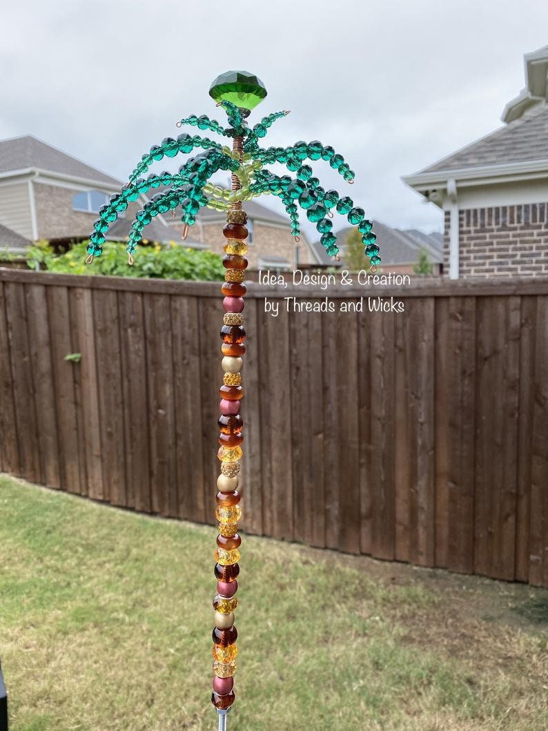 May include: A beaded palm tree decoration with a green crown and a colourful beaded trunk. The beads are in shades of gold, brown, and red. The text "Idea, Design & Creation by Threads and Wicks" is visible on the trunk.