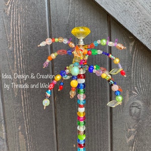 May include: A colourful beaded palm tree garden stake with a yellow glass knob at the top. The stake is made of wire and beads in various colours, including red, orange, yellow, green, blue, purple, and pink. The text "Idea, Design & Creation by Threads and Wicks" is visible at the bottom of the image.