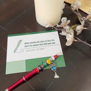 May include: A green card with the text "When writing the story of your life, don't let anyone else hold your pen. Write well, Edit often!"  A red pen with colorful beads and a silver owl charm is on top of the card.