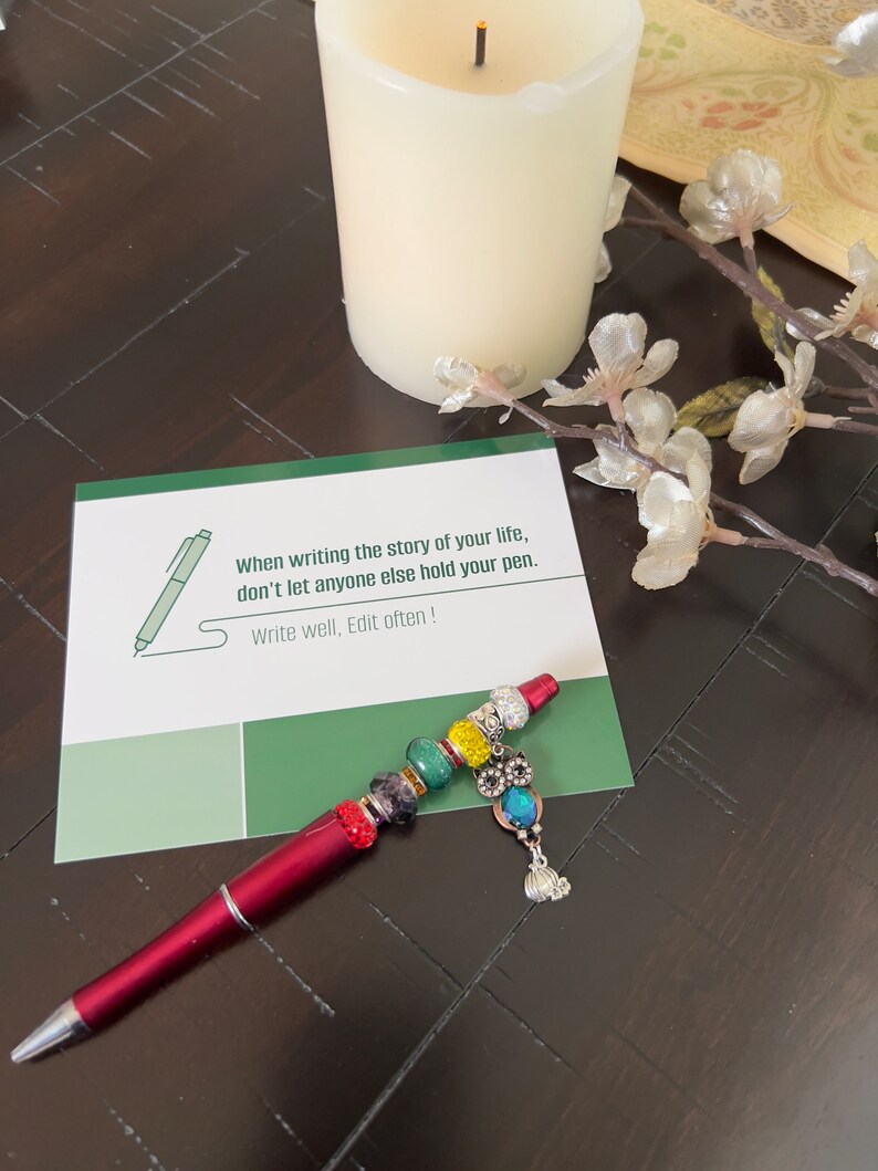 May include: A green card with the text "When writing the story of your life, don't let anyone else hold your pen. Write well, Edit often!"  A red pen with colorful beads and a silver owl charm is on top of the card.