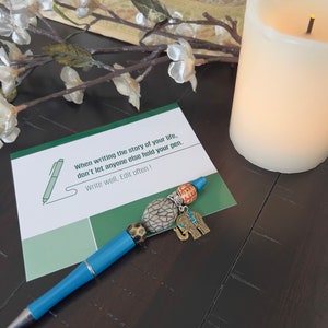May include: A blue pen with a decorative elephant charm and other beads. The pen is on a green card with the text "When writing the story of your life, don't let anyone else hold your pen. Write well, Edit often!"