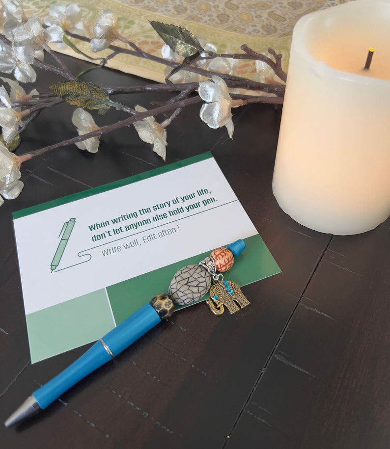 May include: A blue pen with a decorative elephant charm and other beads. The pen is on a green card with the text "When writing the story of your life, don't let anyone else hold your pen. Write well, Edit often!"